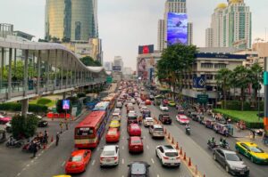 Vista de una avenida principal en Bangkok con mucho tráfico, múltiples taxis coloridos, autobuses y tuk-tuks, rodeados de edificios altos y un paso peatonal elevado lleno de personas.